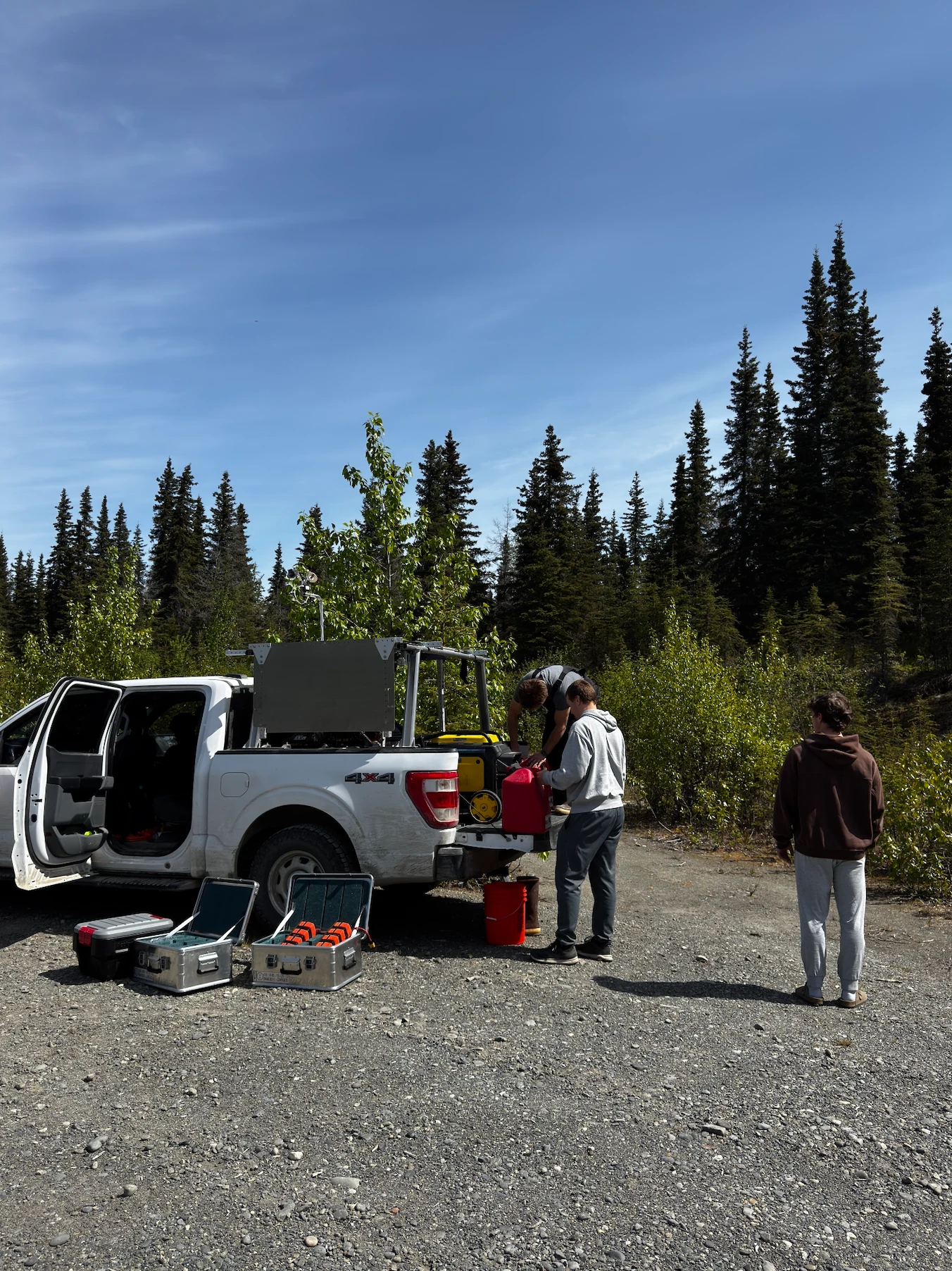 Rainmaker field operators unload equipment from a truck at a gravel staging area in the Alaskan forest.