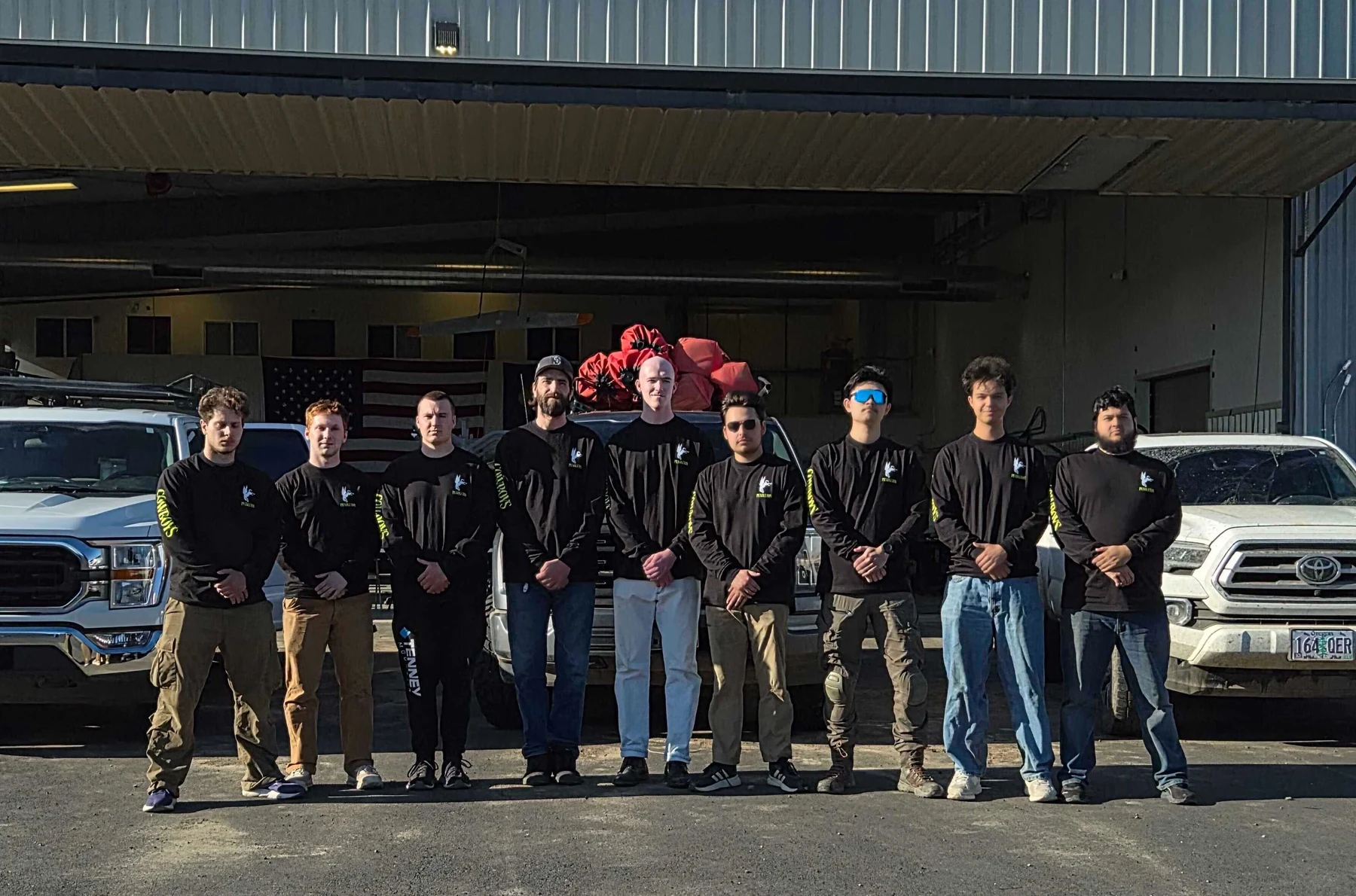 The Rainmaker field crew lines up in front of the hangar between two trucks, an American flag on the wall behind.