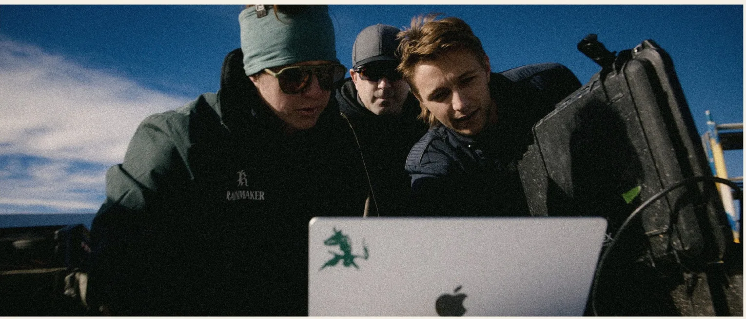 Three operators huddle over a laptop balanced on field cases between storm cells, blue sky breaking behind them.