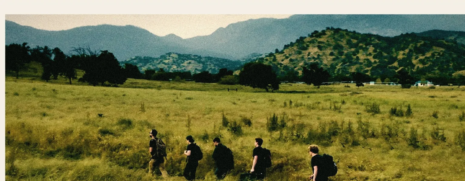 Five crew members with packs walk single file through tall basin grass toward distant mountains.